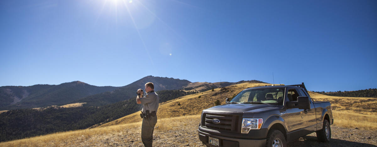 A conservation officer stands next to his truck peering through binoculars.