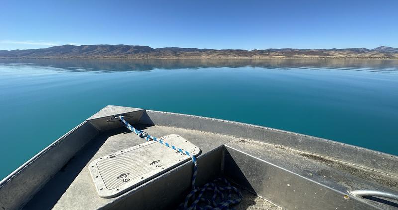 The front of a boat shown atop the bright blue waters of Bear Lake.  Foothills are in the background under a blue sky.